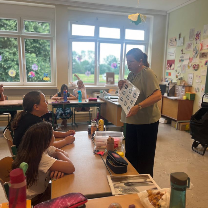 Eine Lehrerin hält ein Stück Papier mit Zahlen in einem Klassenzimmer, während Schüler am Schreibtisch sitzen und ihr zuschauen. Ein Fenster zeigt Bäume draußen.