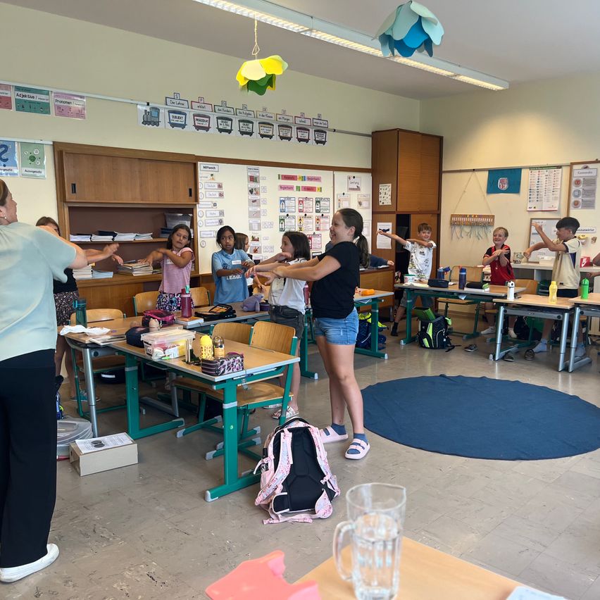 In einem Klassenzimmer stehen Kinder und eine Lehrerin und machen Übungen. Einige Kinder halten eine Flasche. Ein Schreibtisch ist mit verschiedenen Gegenständen gefüllt. Ein Rucksack liegt auf dem Boden.