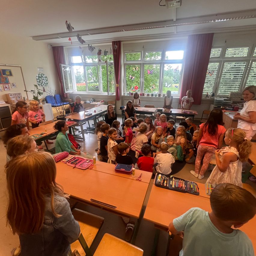 Ein Klassenzimmer mit Kindern und Lehrern, einige sitzen um einen Tisch herum, andere stehen. Es gibt Tische, Stühle und Fenster mit Vorhängen. Draußen sind Bäume zu sehen.