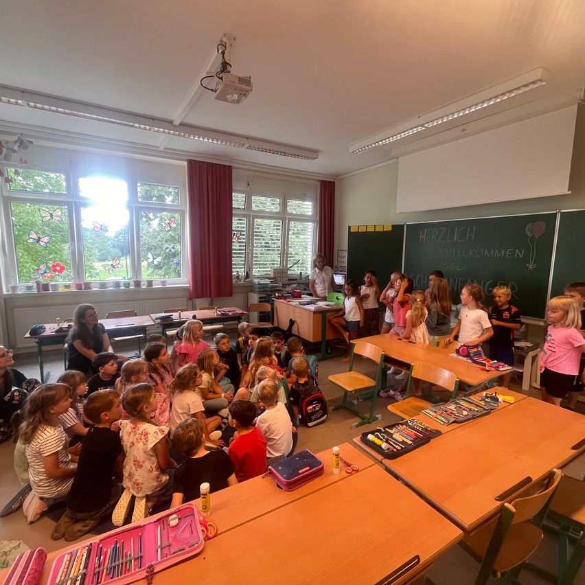 Ein Klassenzimmer voller Kinder, die in einem Kreis sitzen, eine Lehrerin steht in der Nähe der Tafel mit den Worten Herzlich Willkommen darauf geschrieben.