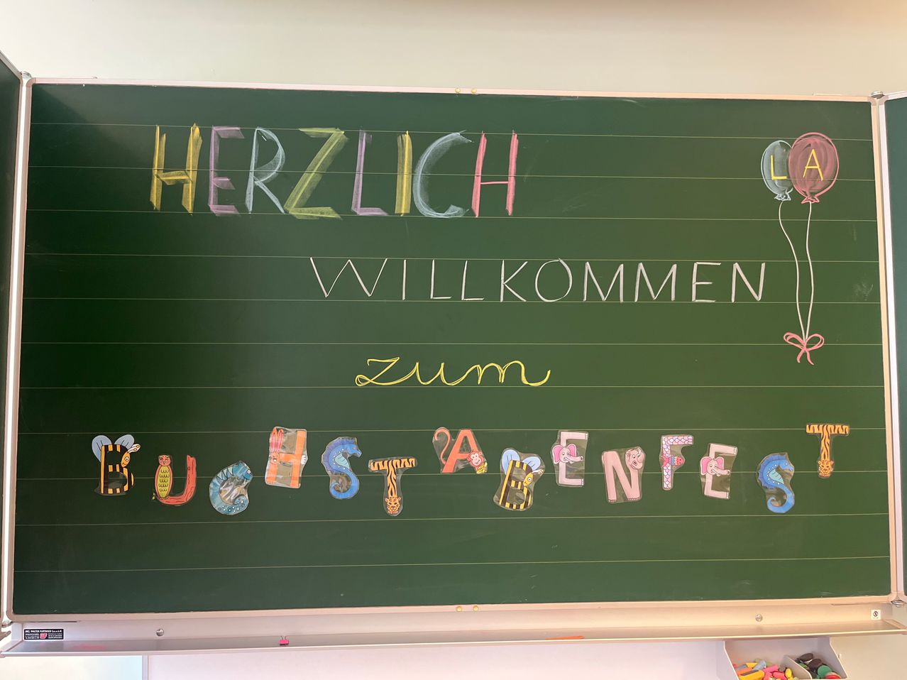 A blackboard with multicolored letters spells out 'Herzlich Willkommen Zum Buchstafenfest'. Below it, colorful letters spell out 'Buchstafenfest' with a balloon in the corner.