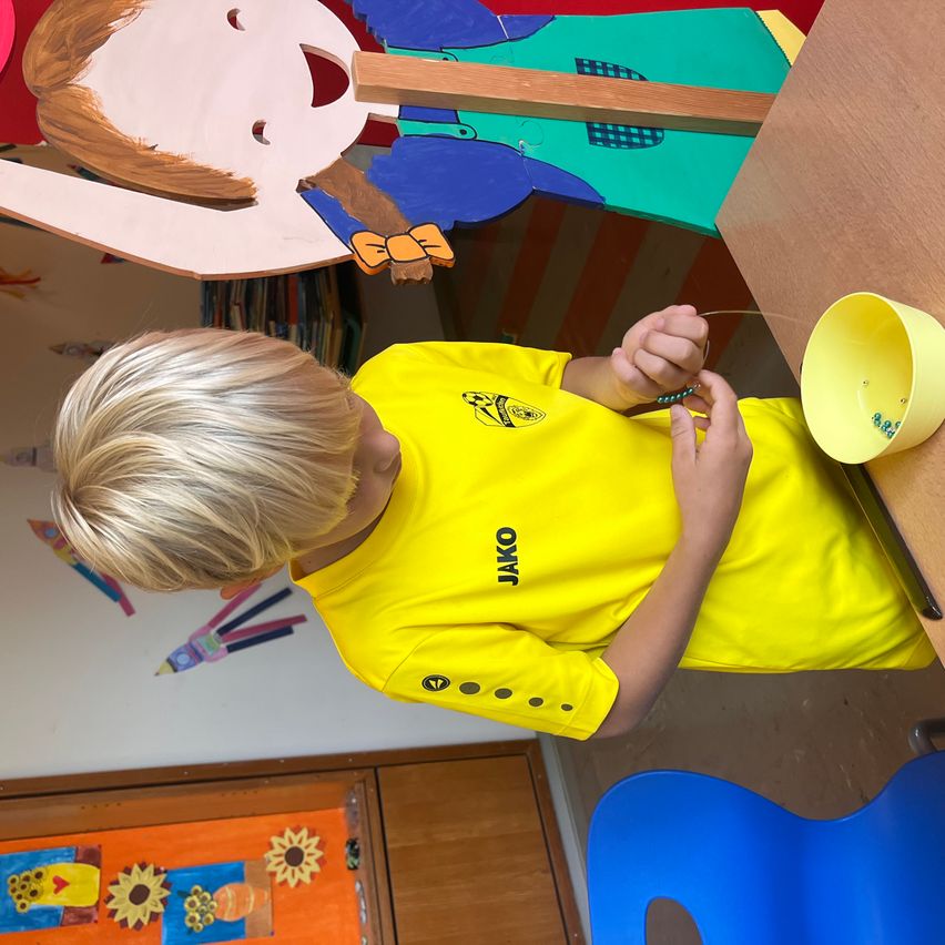 A young boy is wearing a yellow shirt with the word Jako and is creating something with a yellow bucket and a string. He is sitting at a desk with a blue chair.