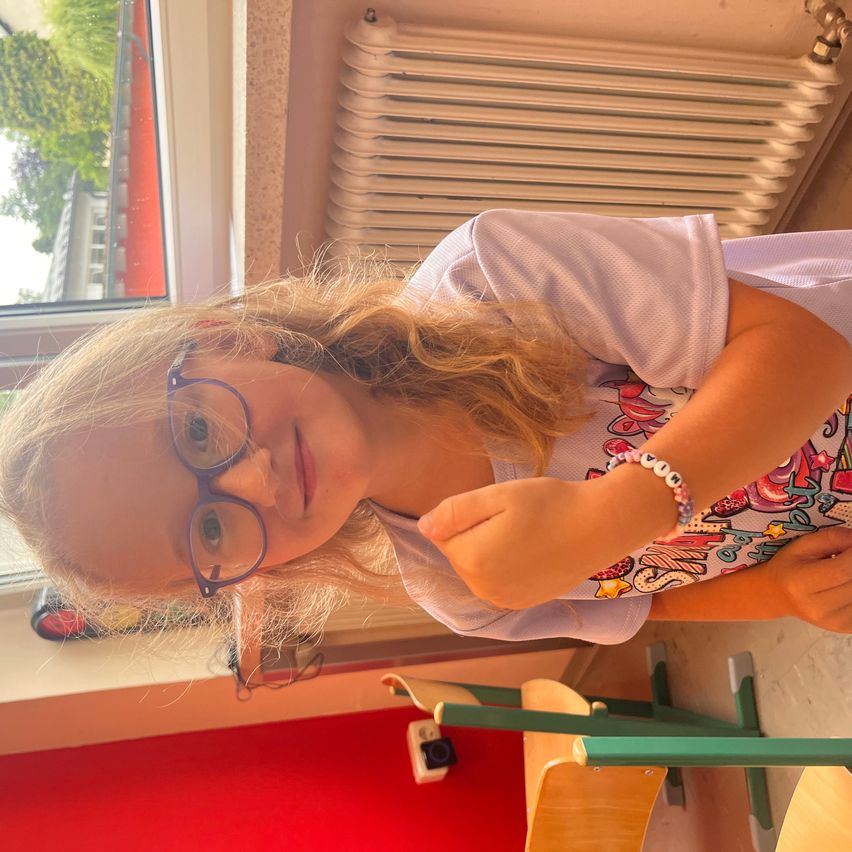 A young girl with glasses and a colorful shirt is smiling and looking at something in a room with a white radiator and a window.