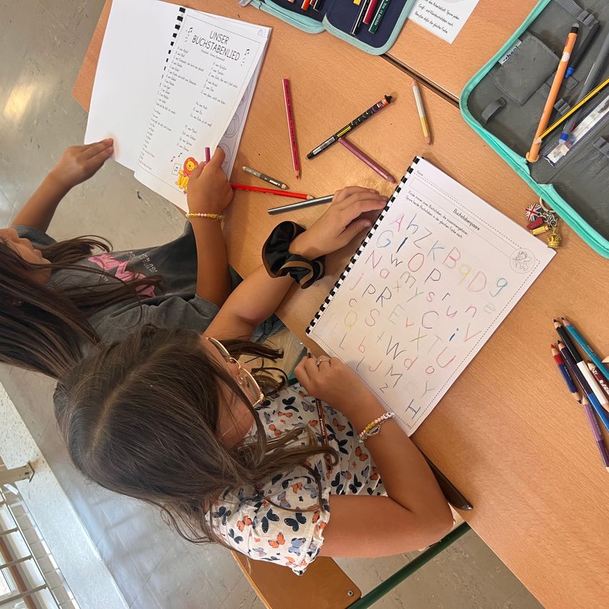 Two children are sitting at a desk, each holding a worksheet. They are writing on the worksheets with markers. A pencil case with colored pencils is also on the desk.