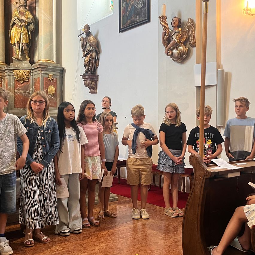 A group of children stand in a church, some holding papers, with religious statues and a red carpet in the background.
