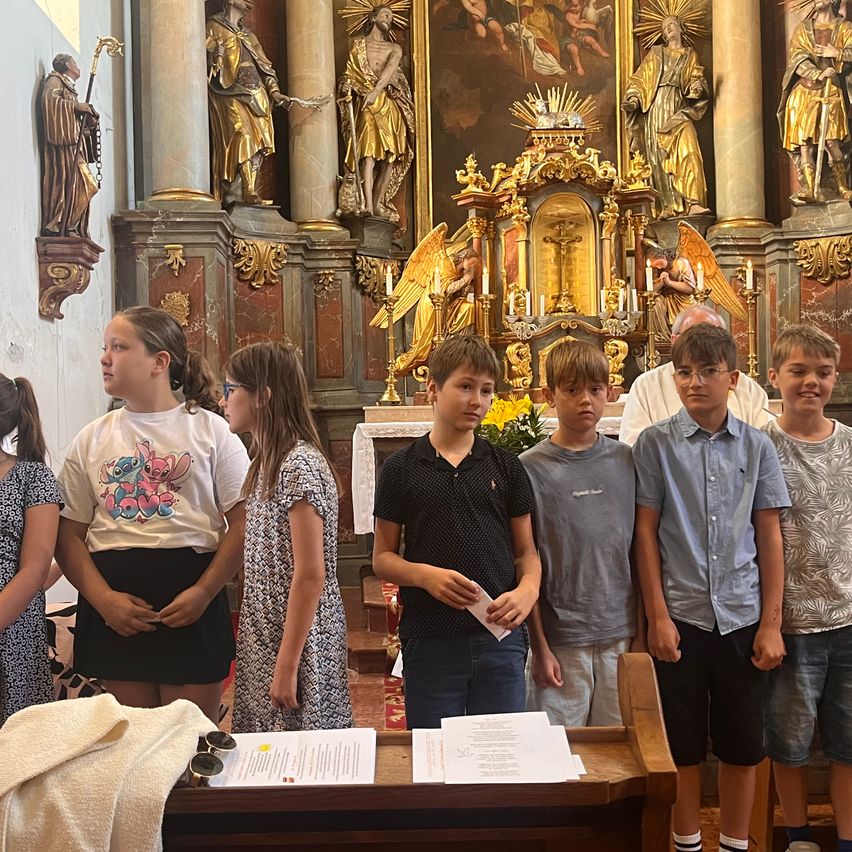 A group of children stands in front of a church altar, with golden statues and a priest behind them. Papers are on the altar, and a towel is on the desk.
