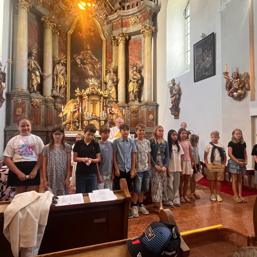 A group of children and adults stand in a church, facing the altar, with a priest in the background. The altar is decorated with statues and a large painting.