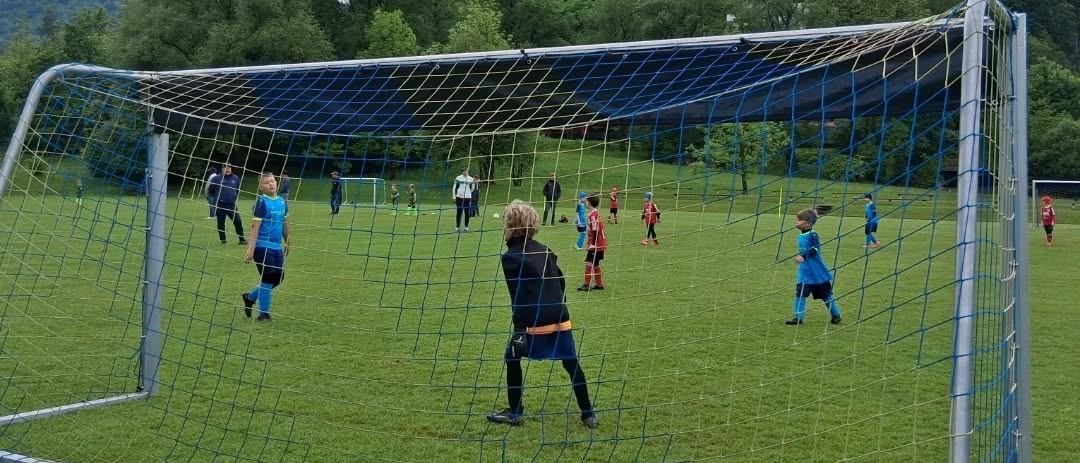 Kinder in blauen Uniformen spielen auf einem Feld Fußball mit einem großen Tor. Bäume und Berge sind im Hintergrund.