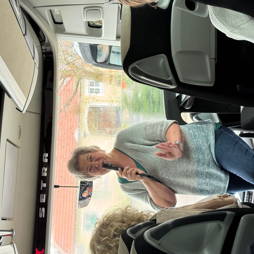 Eine Frau mit einem Mikrofon spricht in einem Fahrzeug, möglicherweise einem Bus, mit einer offenen Tür. Das Fahrzeug hat schwarze Sitze, und das Fenster zeigt ein Haus und Bäume.