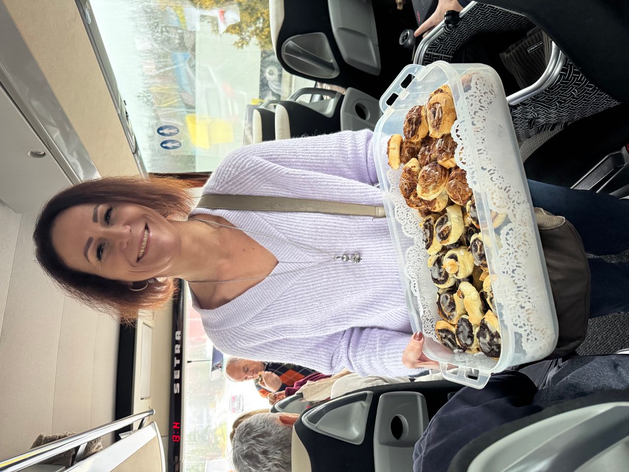 Eine Frau im Bus hält einen Plastikbehälter voller Croissants und lächelt. Hinter ihr sitzen Menschen auf Sitzen, und ein digitales Display zeigt 'Setra' an.