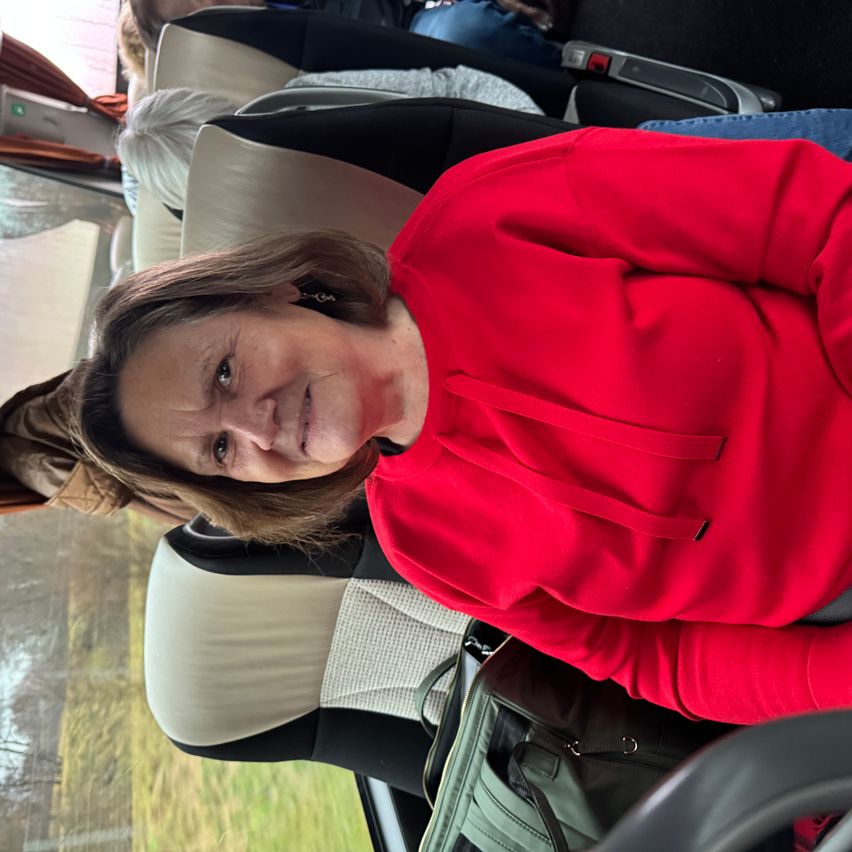 Eine Frau in einem roten Sweatshirt sitzt in einem Bus, lächelt und schaut in die Kamera. Neben ihr sind andere Passagiere, und der Bus fährt auf einer Straße.