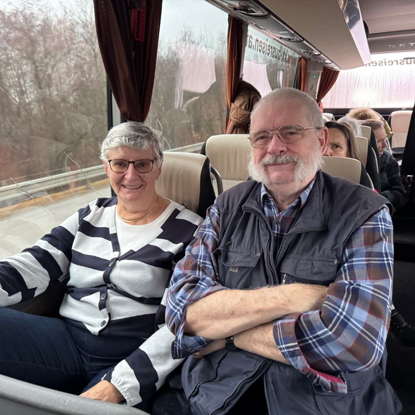 Zwei Personen sitzen in einem Bus, mit Bäumen, die durch das Fenster sichtbar sind. Die Frau trägt eine Brille und eine Halskette. Der Mann hat einen Schnurrbart und einen Bart, trägt eine Brille und eine Weste.