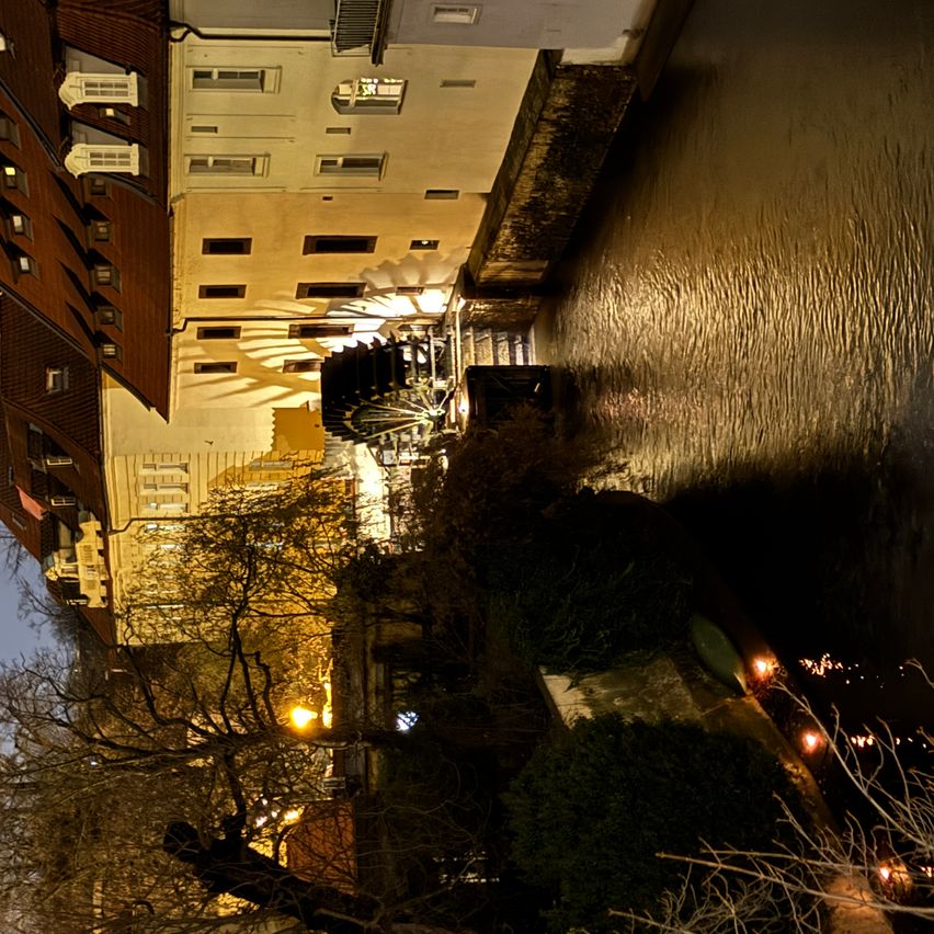 Eine Luftaufnahme eines Kanals mit Gebäuden an der Seite bei Nacht. Bäume und Pflanzen stehen am Kanalrand. Die Gebäude haben viele Fenster und sind von Lichtern beleuchtet.