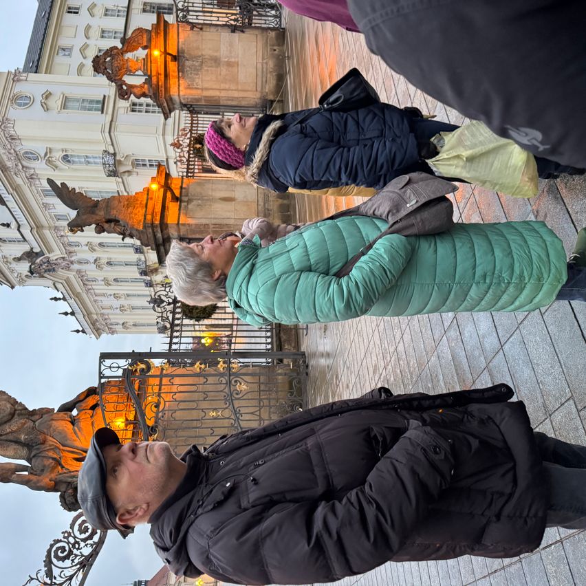 Vier Personen stehen vor einem historischen Gebäude mit Statuen. Zwei Frauen schauen sich etwas an, und ein Mann schaut nach unten. Der Hintergrund ist eine Steinmauer mit einem Metalltor.