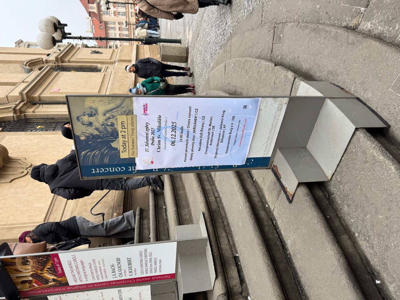 Ein Plakat, das ein Event mit dem Titel "Coro de Navidad" um 14 Uhr ankündigt, mit zusätzlichem Text auf Spanisch. Unter dem Plakat führt eine Treppe zu einem Gebäude hinauf.