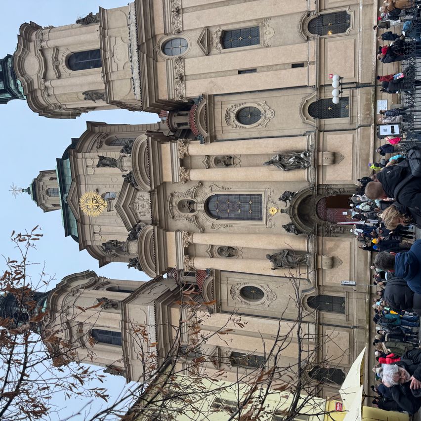 Auf einem Hügel steht eine gotische Kathedrale mit zwei Türmen. Eine Menschenmenge versammelt sich an ihrer Basis unter kahlen Bäumen.