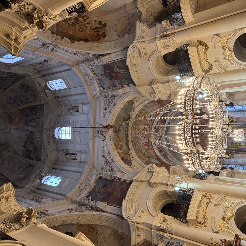 Das Innere einer großen Kirche mit aufwendigen Deckenverzierungen und einer großen Kronleuchter, die von der Mitte hängt. Die Wände und die Kuppel sind mit Gemälden geschmückt.