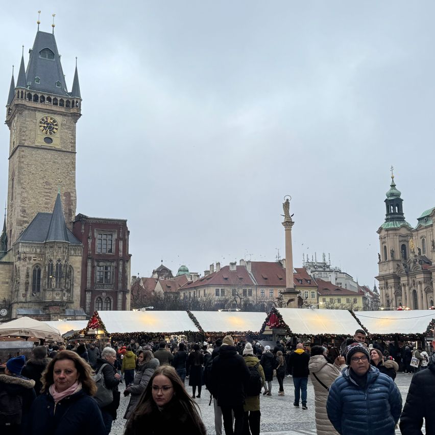 Ein überfüllter Freiluftmarkt auf einem historischen Stadtplatz mit einem großen Uhrturm, einer Statue und Gebäuden. Der Himmel ist bedeckt.