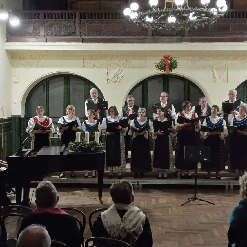 Ein Chor tritt auf einer Bühne mit einem Klavier und einer Kronleuchter darüber auf. Die Mitglieder halten Bücher, und ein festlicher Kranz schmückt die Wand. Zuschauer beobachten von Stühlen aus.
