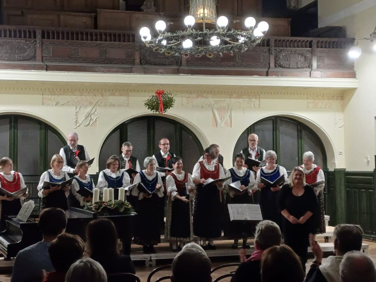 Ein Chor in traditioneller Kleidung steht auf der Bühne und singt, während sie Bücher halten. Dahinter steht ein Klavier, und eine Kronleuchter hängt von der Decke. Mehrere Menschen sitzen im Publikum.
