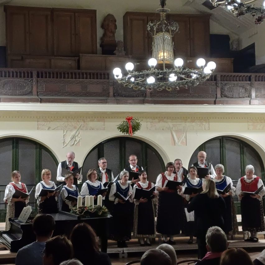 Ein Chor in traditioneller deutscher Kleidung singt auf einer Bühne vor einem Klavier und Kerzen, mit einem Publikum unter einer Kronleuchter.