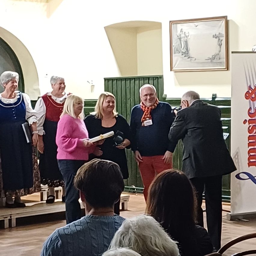 Mehrere Personen, darunter drei Frauen in traditioneller Kleidung, stehen auf einer Bühne und erhalten Auszeichnungen. Ein Mann in Anzug macht ein Foto. Das Publikum schaut von vorne zu.