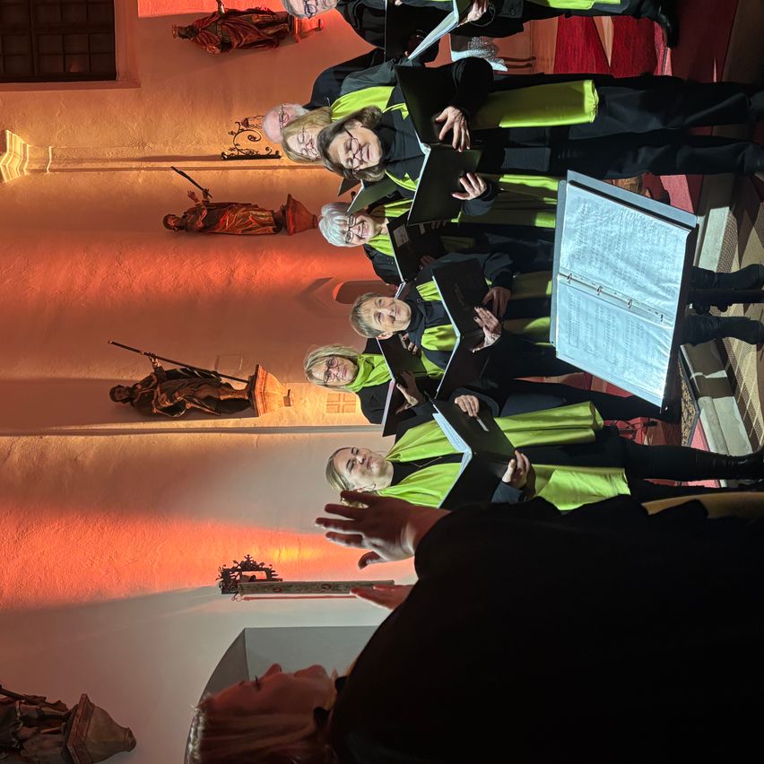 Ein Chor probet in einer Kirche, erhellt von warmer Beleuchtung. Die Mitglieder tragen grüne und schwarze Gewänder und halten Notenbücher. Ein Dirigent steht vorne und dirigiert. Statuen und Ornamente schmücken die Wände.