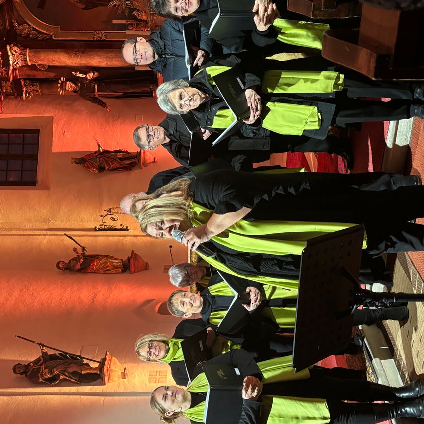 Ein Chor in gelben und schwarzen Gewändern tritt auf, eine Frau hält ein Mikrofon in der Hand und steht vor einer Statue an der Wand.