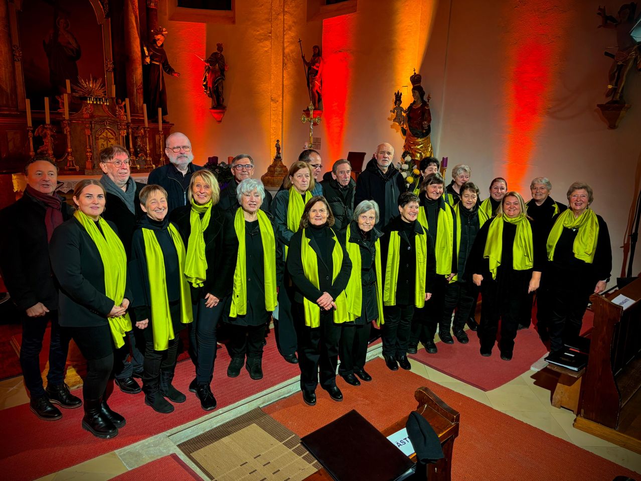 Ein Chor von Menschen in schwarzen und gelben Schals steht in einer Kirche mit beleuchteten Statuen und einem Kreuz im Hintergrund.