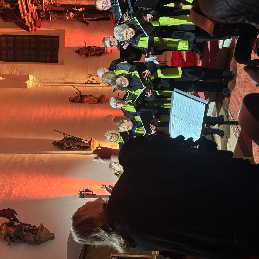 Ein Chor in grünen und schwarzen Westen tritt in einer Kirche auf, an deren Wänden Statuen stehen und ein roter Teppich auf dem Boden liegt.