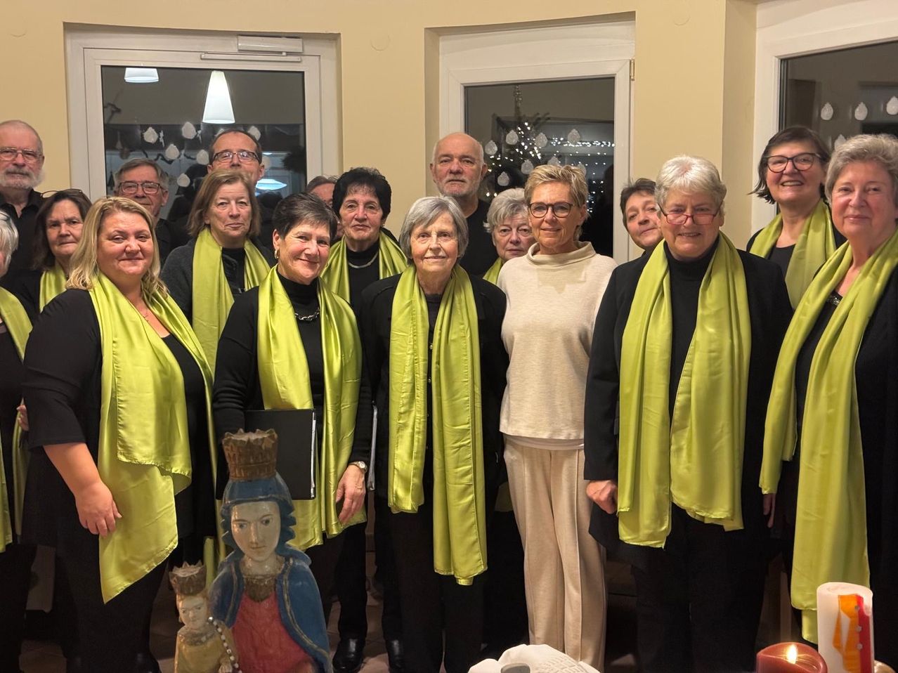 Eine Gruppe von Menschen, die grüne Schals und Brillen tragen, stehen zusammen mit einer Statue in der Mitte. Sie befinden sich in einem Raum mit weißen Türen, Fenstern und Dekorationen.