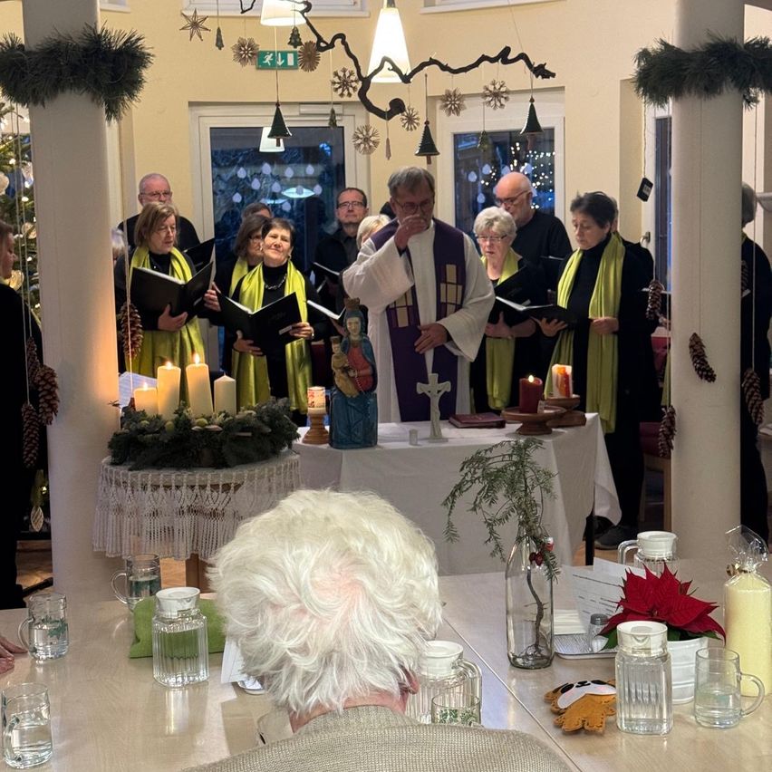 Ein Priester leitet einen Chor während eines Weihnachtsgottesdienstes mit brennenden Kerzen, Dekorationen und einem Weihnachtsbaum im Hintergrund.