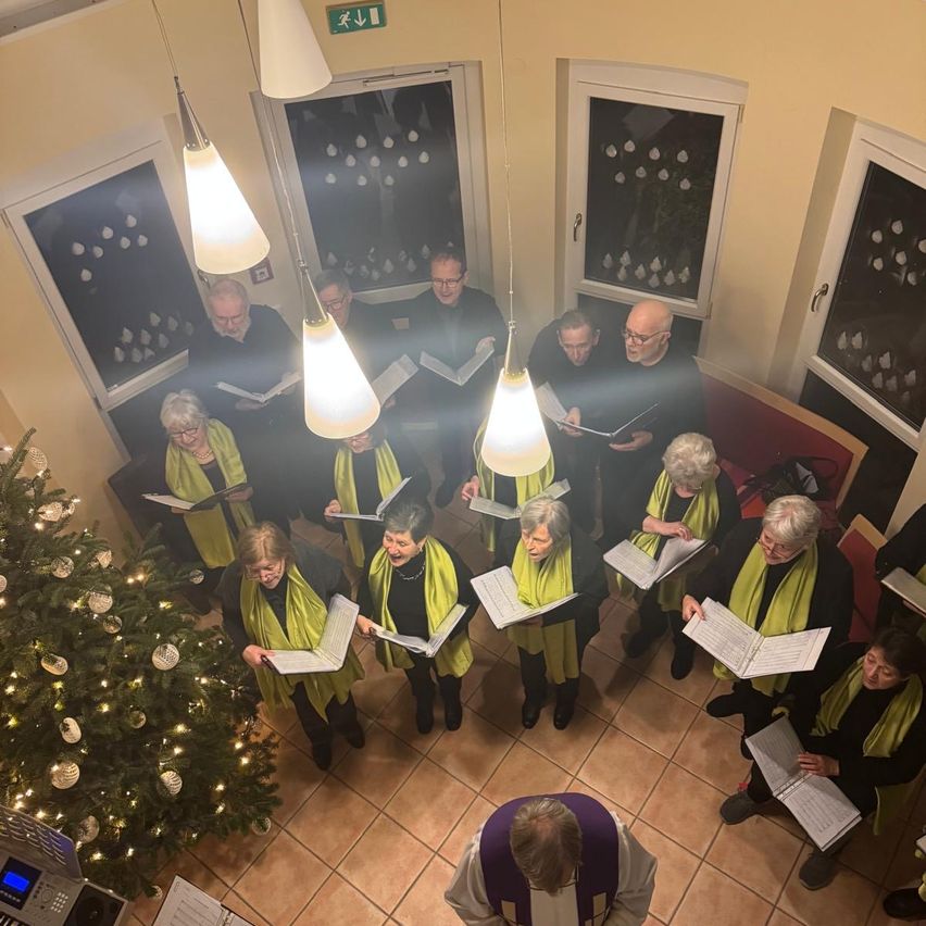 Ein Chor älterer Erwachsener, die grüne Schals tragen, versammelt sich um einen Weihnachtsbaum und singt gemeinsam mit Notenblättern. Der Raum ist warm beleuchtet und der Boden ist gefliest.