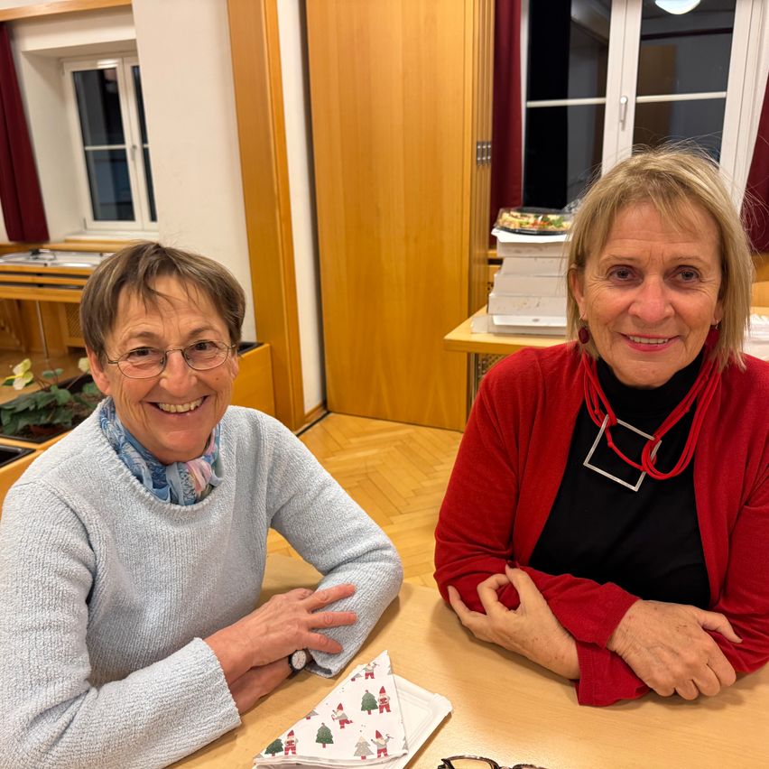 Zwei Frauen sitzen lächelnd an einem Tisch in einem Raum. Eine trägt einen grauen Pullover, die andere einen roten Cardigan über einem schwarzen Top. Ein Weihnachtstuch liegt auf dem Tisch. Hinter ihnen eine Holztür und ein Fenster mit Blick in einen Raum mit einem Schreibtisch.