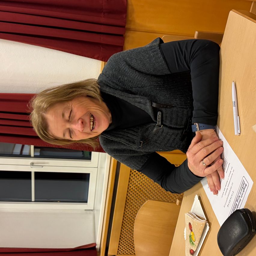 Eine Frau lacht, während sie an einem Tisch sitzt, einen Stift und ein Papier in der Hand hält und eine graue Strickjacke und eine Uhr trägt. Hinter ihr befindet sich ein Fenster mit roten Vorhängen.