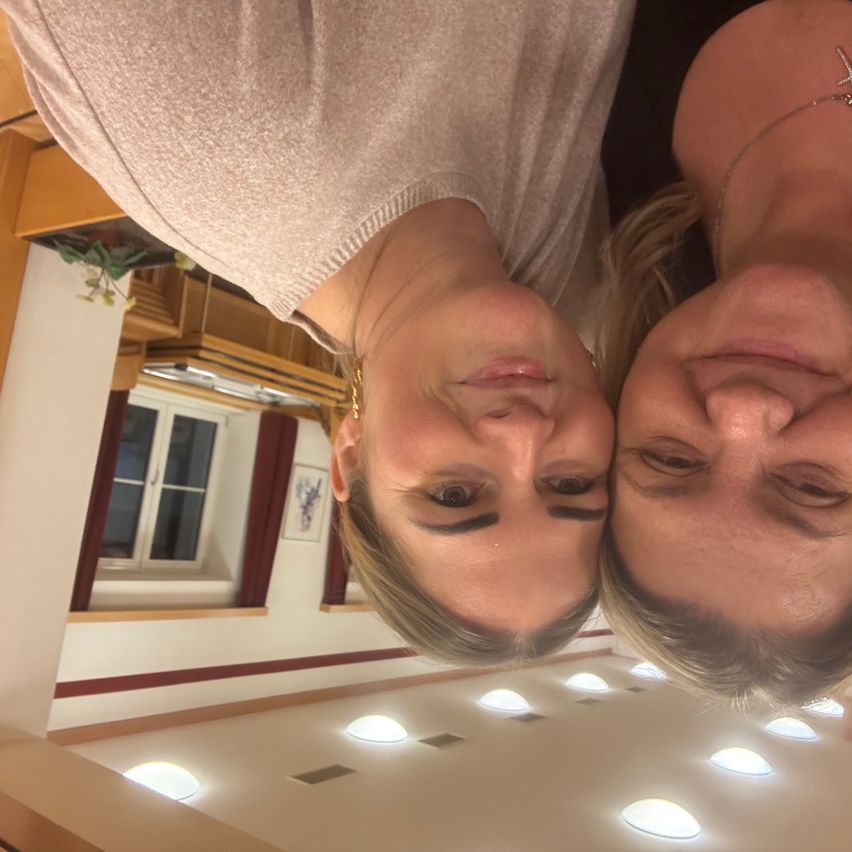 Zwei Frauen sehen sich gegenüber, eine trägt eine Halskette, in einem Raum mit Holzdecken, einem Fenster und roten Vorhängen.