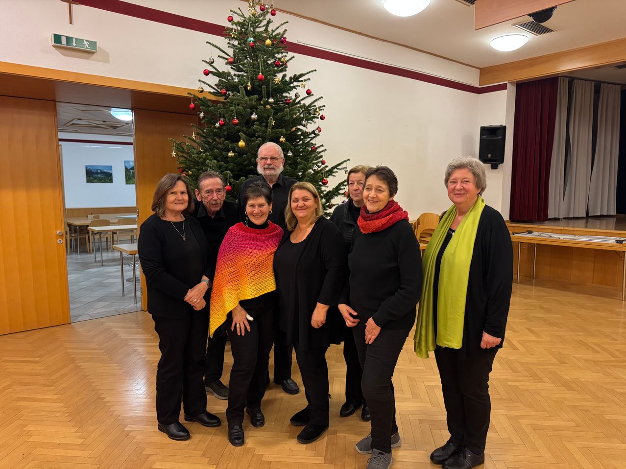 Eine Gruppe von Menschen steht vor einem geschmückten Weihnachtsbaum in einem Raum. Sie tragen schwarze Kleidung und Schals. Der Raum hat einen Holzboden und weiße Wände.