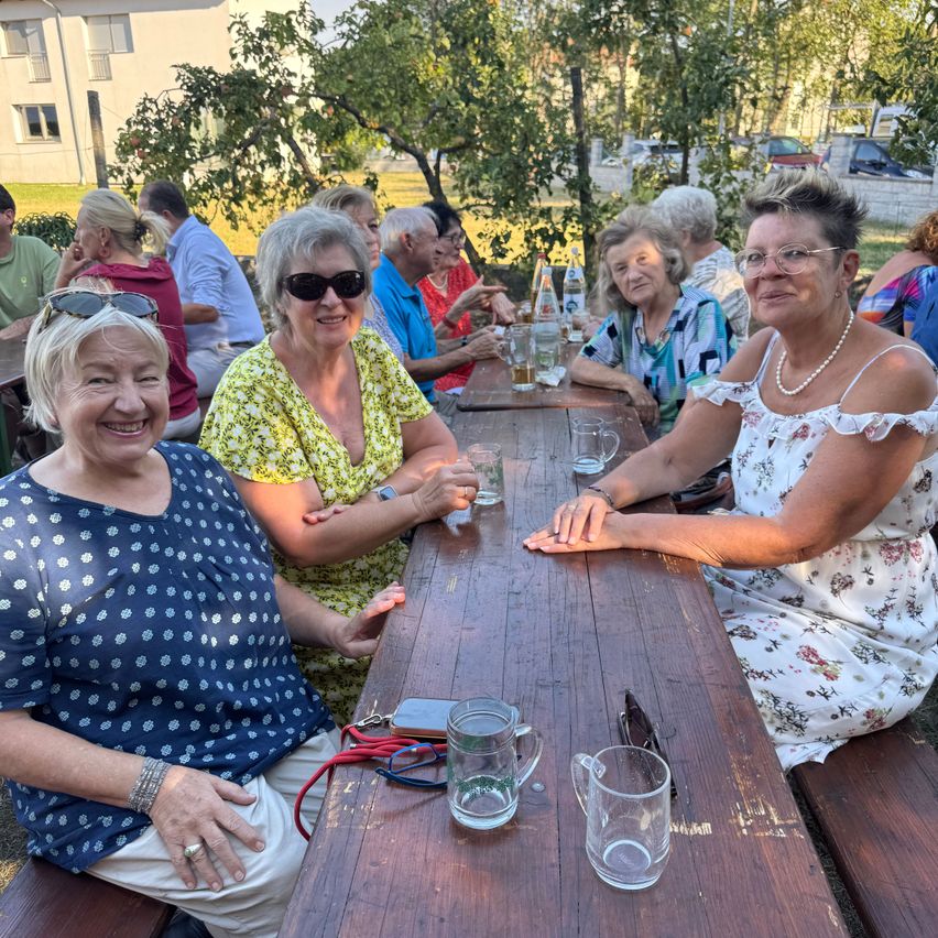 Eine Gruppe älterer Frauen sitzt an einem Holztisch in einer Außenumgebung, lächelt und posiert für ein Foto. Sie sind von anderen Leuten umgeben, die auf Bänken sitzen, mit Bäumen und Gebäuden im Hintergrund.
