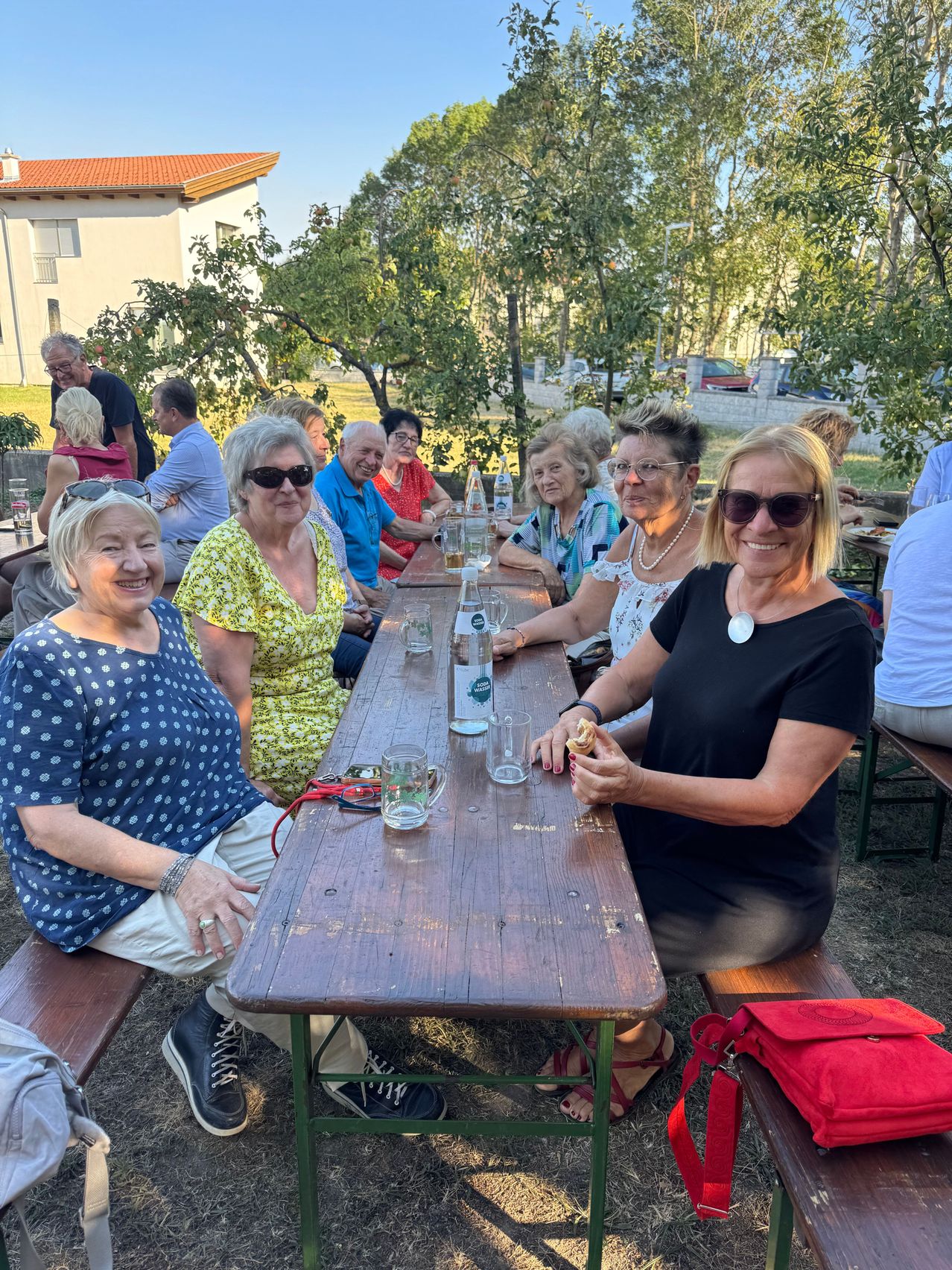 Eine Gruppe von Menschen sitzt an einem langen Holztisch in einer Außenumgebung. Sie lächeln und scheinen eine Mahlzeit gemeinsam zu genießen. Auf dem Tisch stehen Gläser, Flaschen und eine rote Tasche. Bäume und ein Gebäude sind im Hintergrund zu sehen.