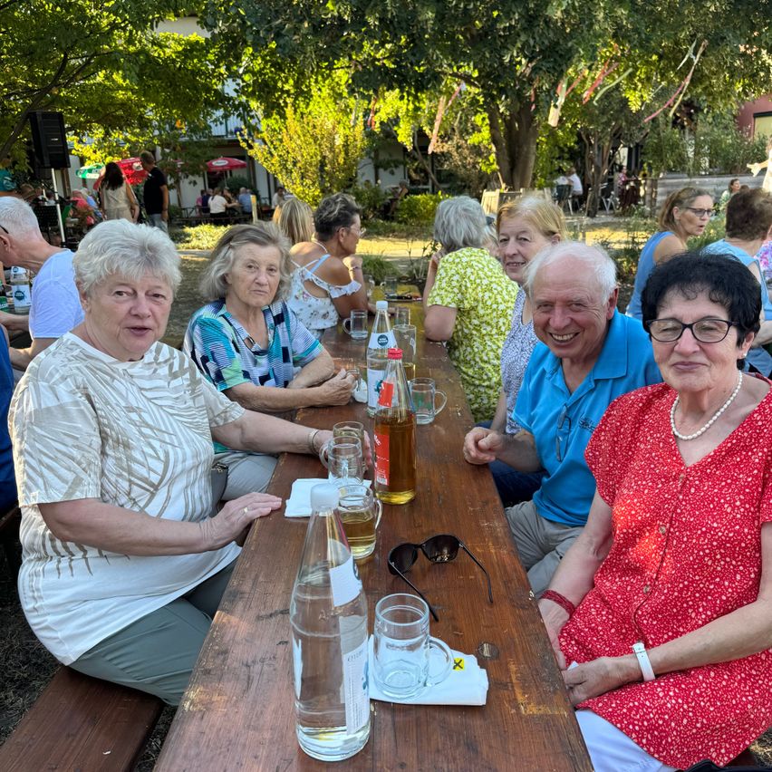 Eine Gruppe älterer Menschen sitzt an einem langen Tisch in einem Garten im Freien und posiert für ein Foto. Sie sind von Flaschen, Gläsern und anderen Gegenständen umgeben. Bäume und Pflanzen sind im Hintergrund.