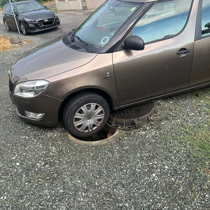 Ein brauner Skoda-Wagen ist teilweise in einem Schachtdeckel in einem Schotterparkplatz versunken. Ein schwarzer Wagen ist dahinter geparkt.