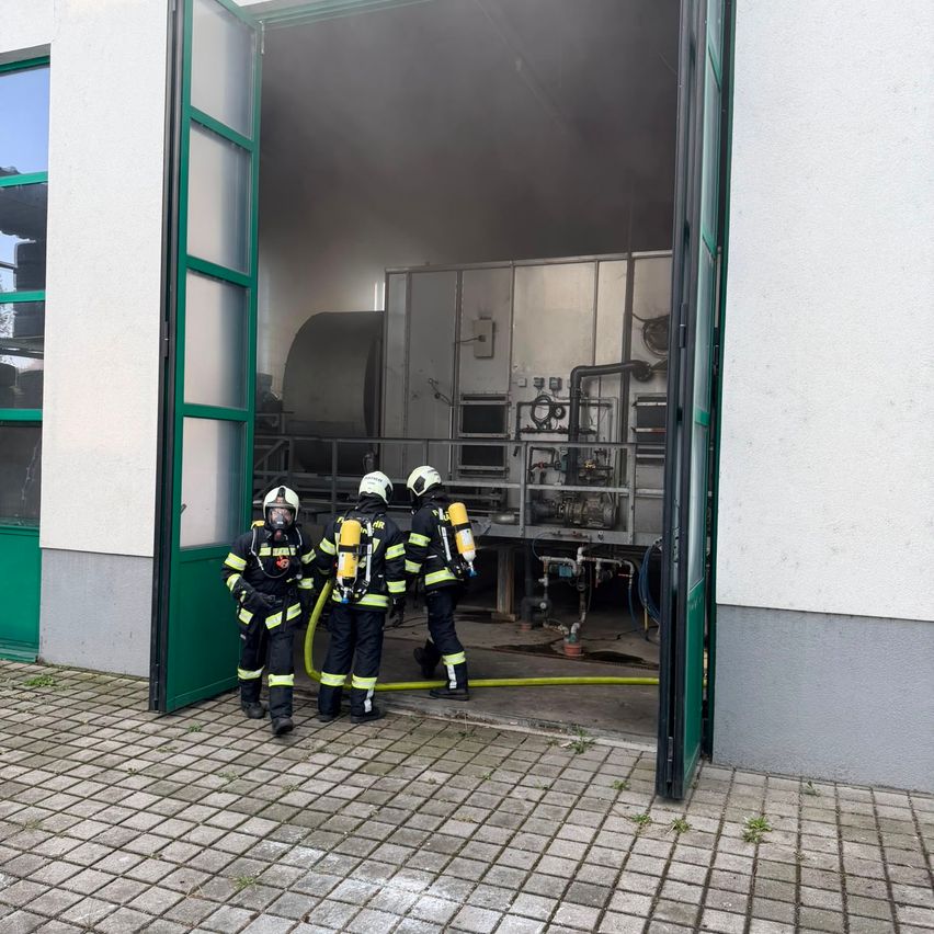 Drei Feuerwehrleute in voller Ausrüstung stehen in der Tür eines Gebäudes, möglicherweise in Vorbereitung auf einen Notfall. Das Innere zeigt Maschinen und Ausrüstung.