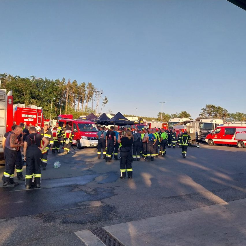 Eine Gruppe von Feuerwehrleuten in Uniform versammelt sich im Freien, mit Feuerwehrwagen und Zelten im Hintergrund.