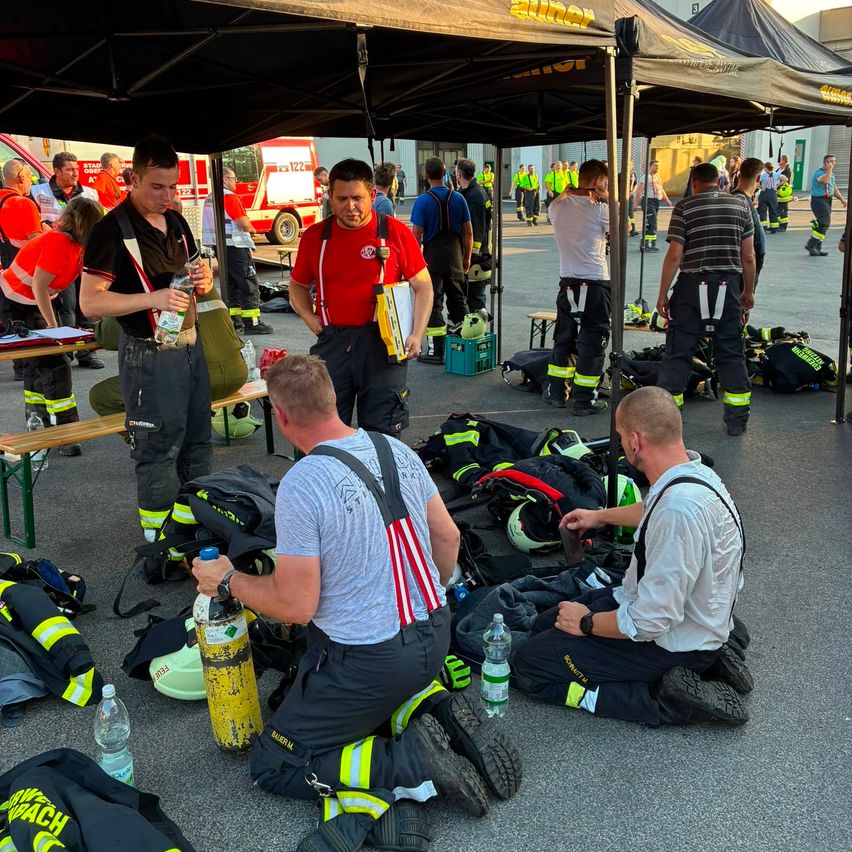 Feuerwehrleute in Uniform versammeln sich unter einem Zelt, einige sitzen und andere stehen. Ausrüstung und Ausrüstungsgegenstände sind verstreut. Ein roter Krankenwagen ist in der Nähe geparkt.
