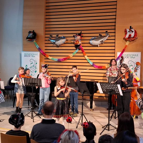 Eine Gruppe von Kindern, die Geige spielen auf der Bühne, während ein Publikum zusieht, begleitet von zwei Erwachsenen, mit Luftballons, die darüber hängen.