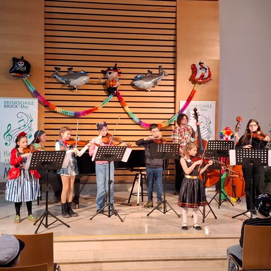 Kinder in Kostümen spielen ein musikalisches Konzert auf der Bühne, mit Violinen, Celli und einem Klavier. Es gibt bunte Dekorationen und Luftballons, die an der Wand hängen.