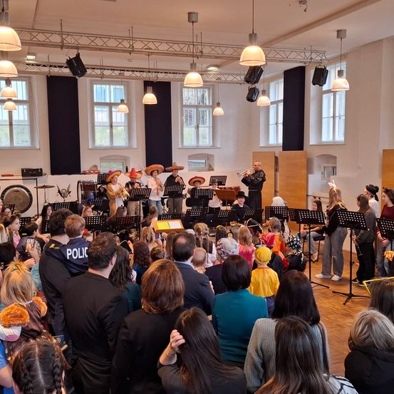 Eine große Gruppe von Menschen ist in einem Saal versammelt, um eine musikalische Aufführung zu verfolgen. Mehrere Musiker, die Hüte tragen, spielen Instrumente, und das Publikum, darunter auch Kinder, schaut zu. Der Saal hat hohe Decken mit hängenden Lampen und großen Fenstern.