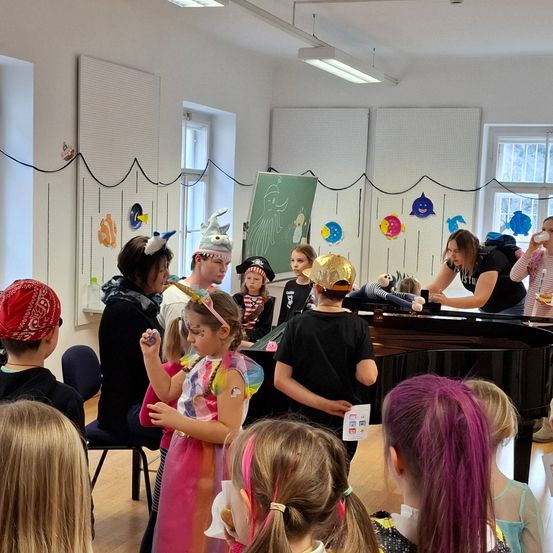 Eine Gruppe von Kindern und Erwachsenen versammelt sich in einem Raum. Sie tragen Kostüme und einige halten Requisiten. Eine Frau steht an einem Klavier.