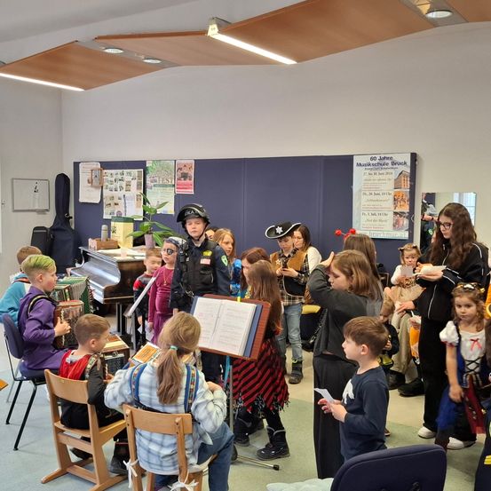 In einem Klassenzimmer versammeln sich Kinder in Kostümen um einen Polizeibeamten und eine Frau, möglicherweise für eine Aufführung oder an einer thematischen Veranstaltung teilnehmend.