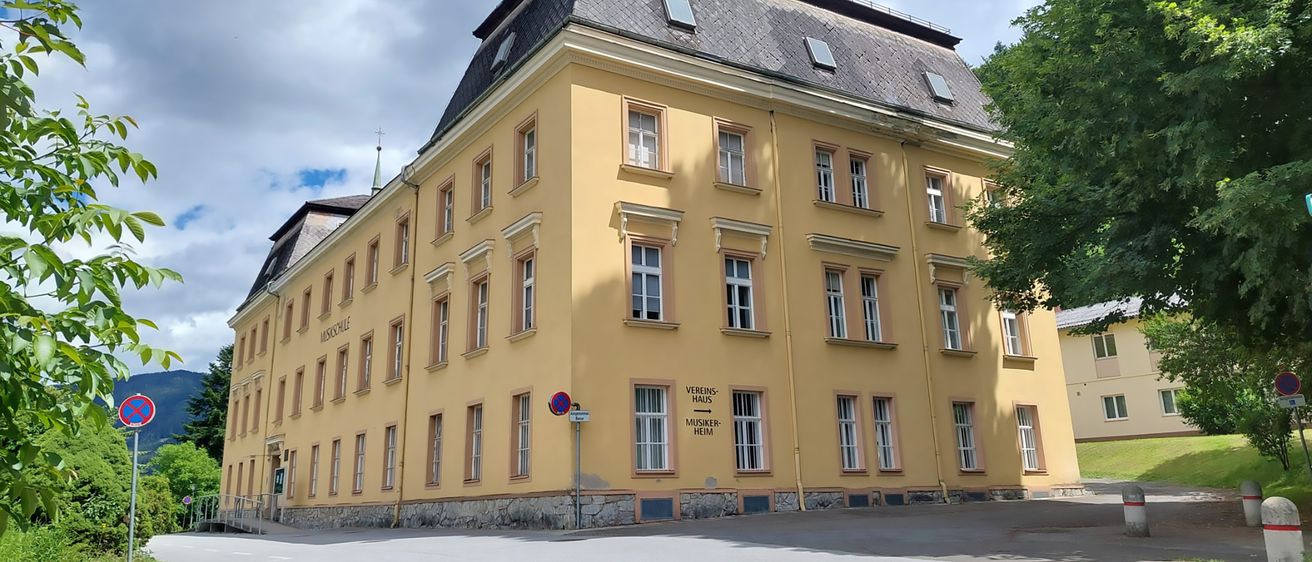 Ein gelbes Gebäude mit vielen Fenstern und einem grauen Dach, beschriftet mit 'Vereins Haus Musikheim', steht unter einem blauen Himmel mit weißen Wolken.